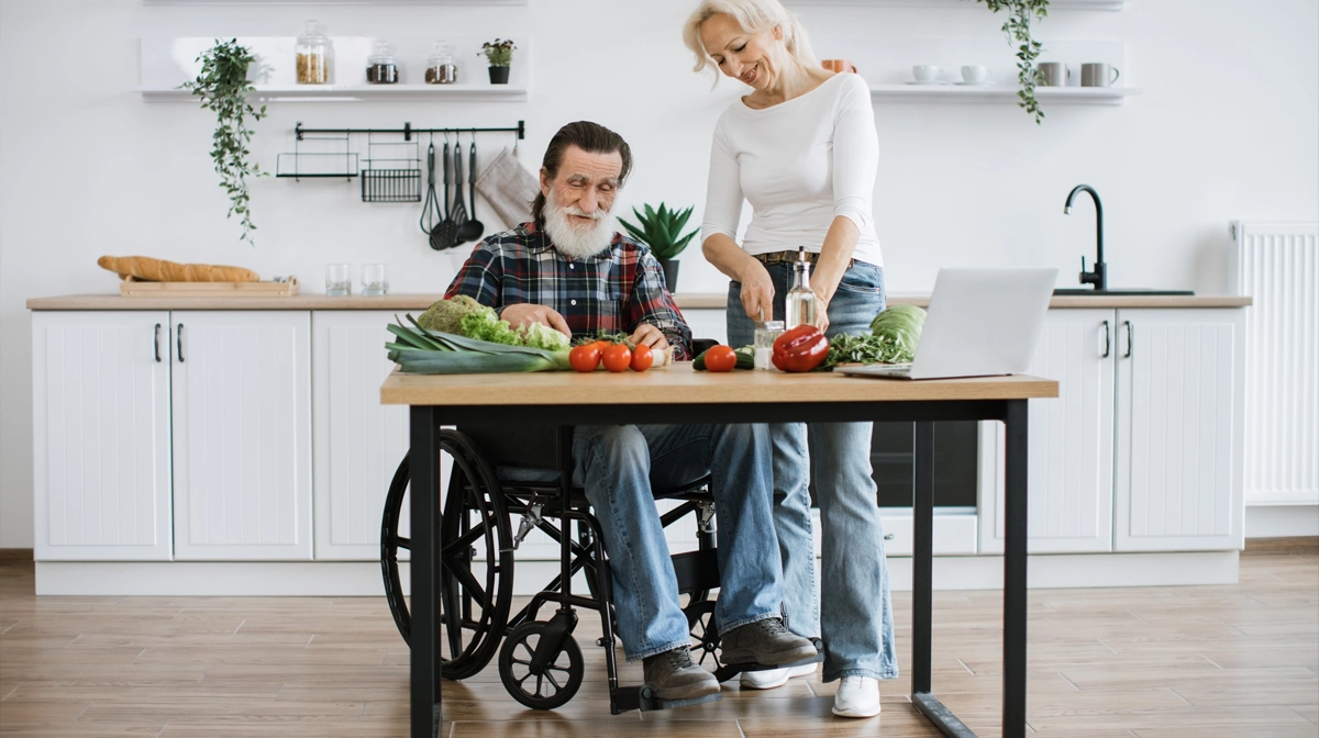 senior man in wheel chair with woman doing kitchen chores